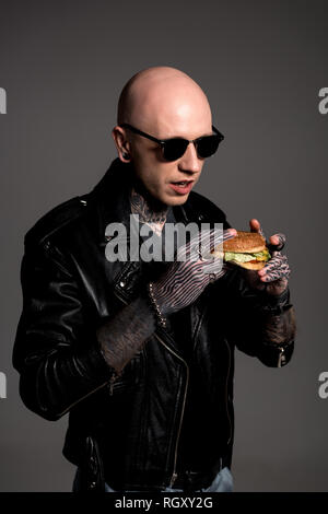 bald tattooed man in leather jacket and sunglasses holding tasty burger isolated on grey Stock Photo