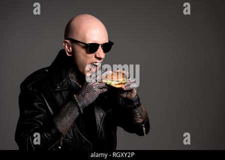 bald tattooed man in leather jacket and sunglasses eating burger isolated on grey Stock Photo