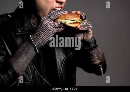 cropped shot of stylish tattooed man in leather jacket eating burger isolated on grey Stock Photo