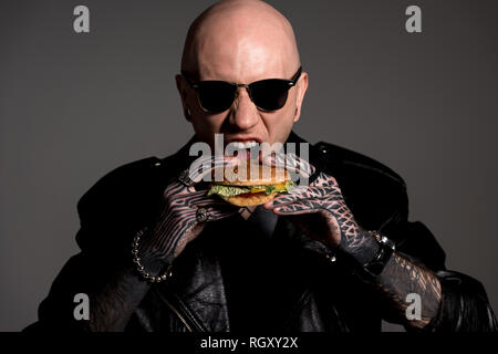 hungry tattooed man in leather jacket and sunglasses eating hamburger isolated on grey Stock Photo
