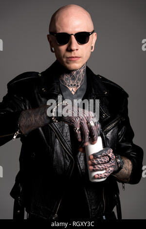 bald tattooed man in leather jacket and sunglasses opening soda can isolated on grey Stock Photo