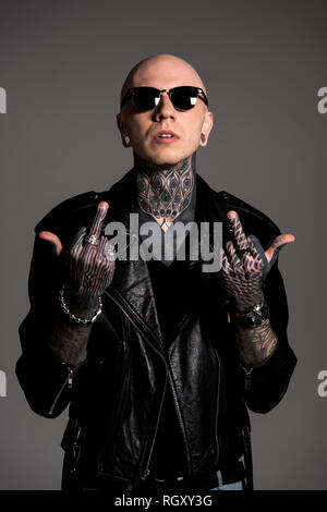 bald tattooed man in leather jacket and sunglasses showing middle fingers isolated on grey Stock Photo