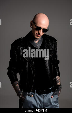 smiling bald tattooed man in leather jacket and sunglasses standing with hands in pockets isolated on grey Stock Photo