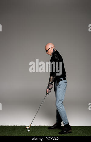 side view of bald tattooed man in leather jacket and sunglasses playing golf on grey Stock Photo