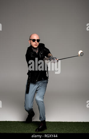 bald tattooed man in leather jacket and sunglasses playing golf on grey Stock Photo