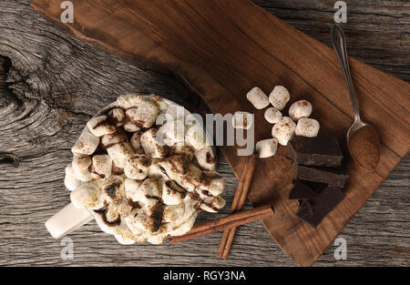 Overhead shot of a mug of hot cocoa with toasted marshmallows next to a cutting board with chocolate chunks and cinnamon sticks. Stock Photo
