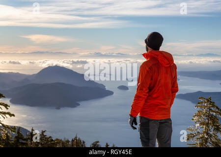 Adventurous man enjoying the beautiful view on top of a mountain during a winter sunset. Taken on Mnt Harvey, near Vancouver, BC, Canada. Stock Photo