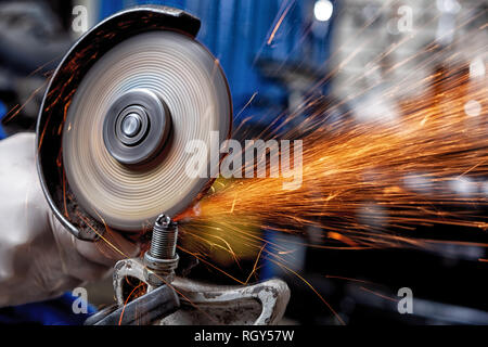 Close-up on the sides fly bright sparks from the angle grinder machine. A young male welder in a white working gloves grinds a metal product with angl Stock Photo