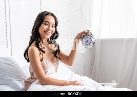 Smiling beautiful asian woman holding alarm clock while sitting in bed and looking at camera Stock Photo