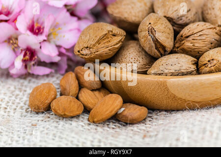 Almonds with shell in wooden bowl, almonds spilled on sack surface, with almond flowers background Stock Photo