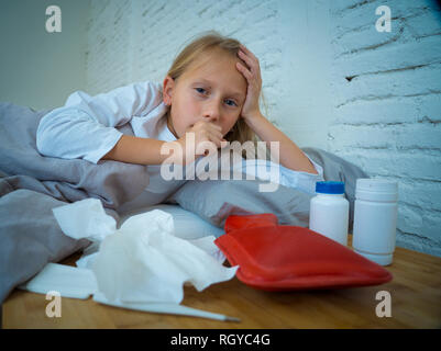 Sweet sick cute girl feeling sick lying in bed with medicines thermometer hot water bag suffering from Cold and Winter Flu Virus Sneezing Running Nose Stock Photo