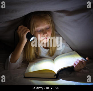 Beautiful caucasian girl lying in bed under the duvet holding a lantern reading a mystery book in the dark late at night looking frightened with a dra Stock Photo