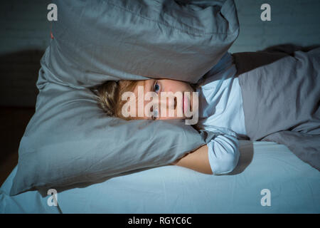 Unhappy girl covering her ears with pillow trying to sleep at night feeling sleepless sad and tired in Parents arguing Family fights Children Sleeping Stock Photo