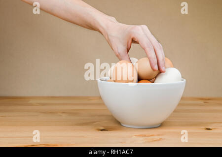 Female hand taking one egg from white bowl on a table . Stock Photo