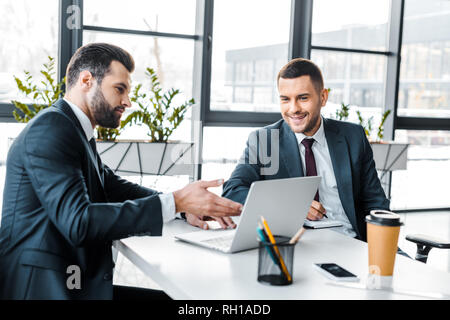 handsome businessman showing something on laptop to smiling coworker in modern office Stock Photo
