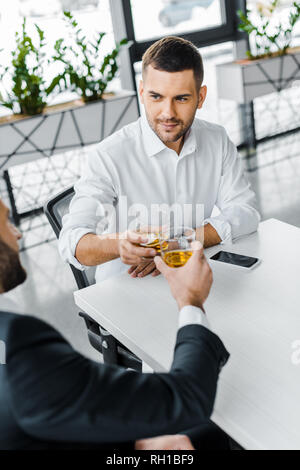 handsome businessman toasting with whiskey while sitting in modern office with coworker Stock Photo