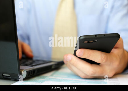 Man in blue shirt and tie works in office, sitting at a table with laptop and smartphone in hand. Concept of clerk, manager or bank employee Stock Photo