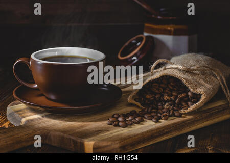 Steaming cup of coffee with coffee beans spilled from a burlap bag on a wooden board in a dark rustic setting Stock Photo