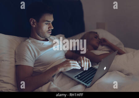 suspicious young man using laptop while girlfriend sleeping in bed at night Stock Photo