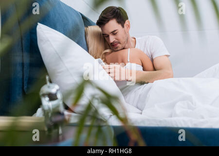 selective focus of smiling young man using smartphone while lying with girlfriend in bed Stock Photo