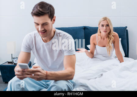 smiling young man in pajamas using smartphone and angry girlfriend looking at camera in bedroom, mistrust concept Stock Photo