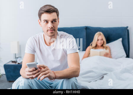 young man holding smartphone and looking away while girlfriend reading book in bed behind Stock Photo