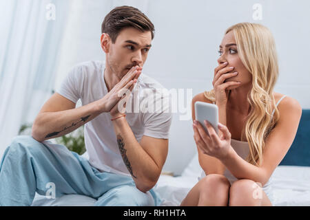 shocked woman holding smartphone and looking at confused husband sitting on bed, mistrust concept Stock Photo