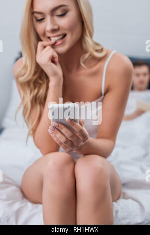 smiling young woman biting nail and using smartphone while husband lying in bed behind Stock Photo