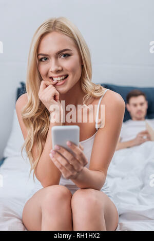 young woman with smartphone biting nail and smiling at camera while husband reading book on bed Stock Photo