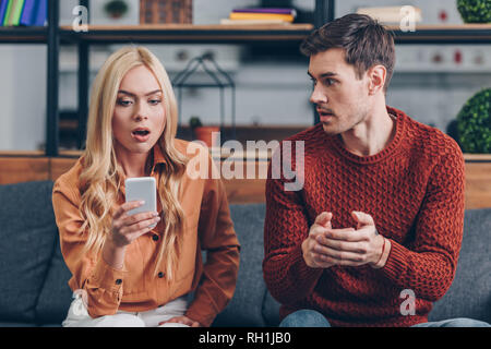 emotional young man looking at shocked girlfriend using smartphone on couch, jealousy concept Stock Photo