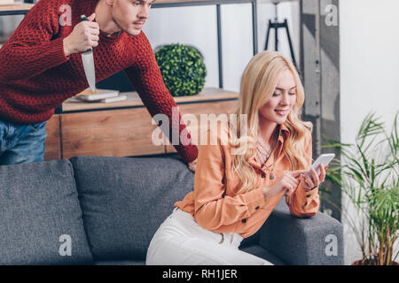 cropped shot of aggressive man with knife looking at smiling wife using smartphone on couch, jealousy concept Stock Photo