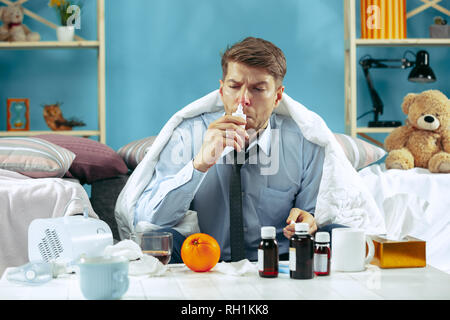 Bearded sick man with flue sitting on sofa at home covered with warm blanket and using nasal spray. The illness, influenza, pain concept. Relaxation at Home. Healthcare Concepts. Stock Photo