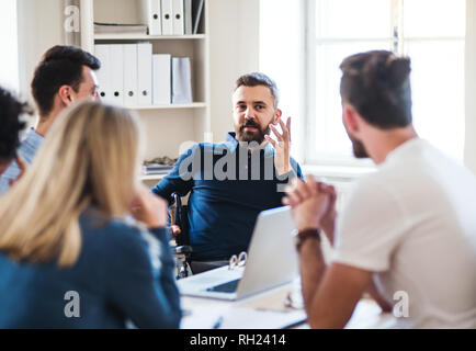 A businessman in wheelchair with colleagues sitting around table, working in a modern office. Stock Photo