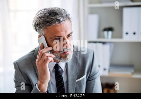 Serious mature businessman with smartphone sitting at the table, making a phone call. Stock Photo