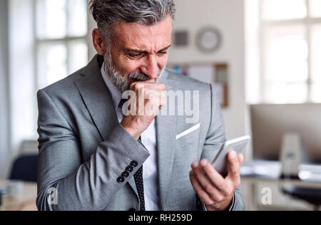A frustrated businessman with smartphone standing in an office, reading bad news. Stock Photo