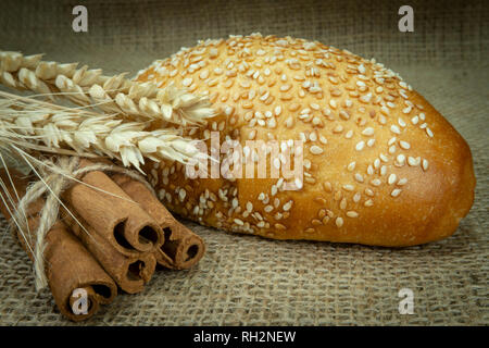 Single sesame seed bun, ear of ripe wheat and bundle of cinnamon sticks on jute fabric surface Stock Photo