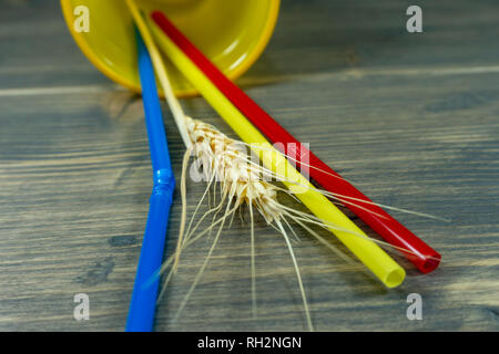 Three colored plastic straw lying on their sides in a yellow beaker with an ear of ripe golden wheat on wood Stock Photo
