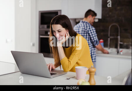 A young attractive woman using laptop in a kitchen and a man washing the dishes. Stock Photo