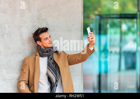 Side view of young trendy man leaning on a wall outdoors wearing denim clothes while taking a selfie in sunny day Stock Photo