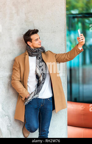 Side view of young trendy man leaning on a wall outdoors wearing denim clothes while taking a selfie in sunny day Stock Photo