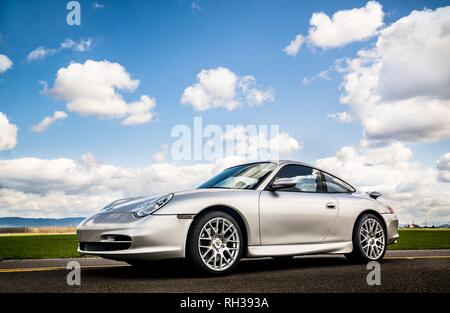 April 2, 2018 Eugene Oregon - A silver Porsche 911 body style 996 sits on an empty road under a sunny cloud filled sky near some green fields of grass Stock Photo