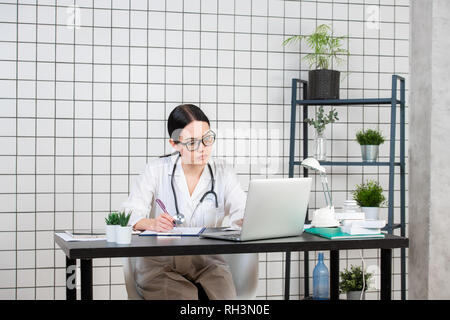 Beautiful female doctor writing while using laptop at desk in clinic. Stock Photo