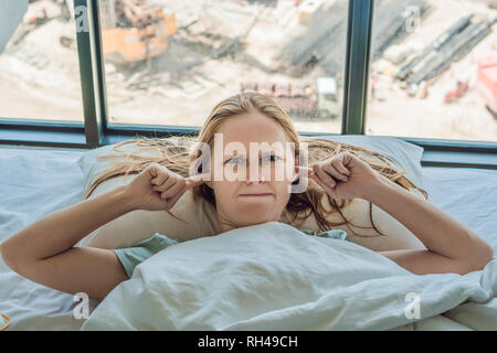 Young woman lying on a bed covered her ears because of the noise. In the window after the bed you can see the construction of a new house Stock Photo