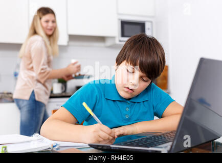 Positive schoolboy doing homework using laptop at kitchen interior, woman cooking at background Stock Photo