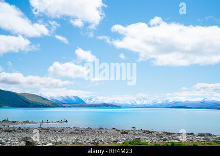 LAKE TEKAPO NEW ZEALAND - OCTOBER 14 2018;Expansive views turquoise water of Lake Tekapo with snow-capped Southern Alps behind and tourists on rocky f Stock Photo