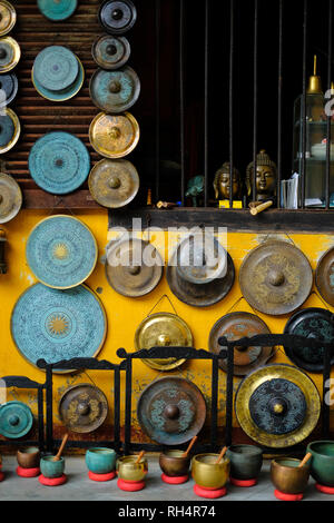 A colorful collection of ornamental gongs, sound bowls and buddha statues hanging on a yellow wall in a shop front in Hoi An, Vietnam. Stock Photo