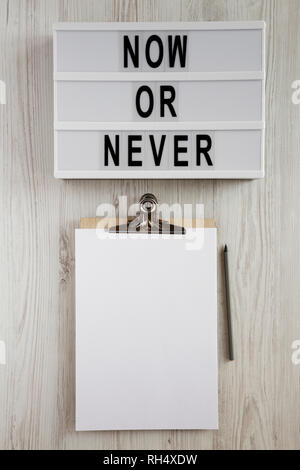 'Now or never' word on modern board, clipboard with sheet of paper on a white wooden background, top view. Space for text. Stock Photo