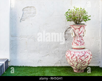 Big vintage white vase with plant on green lawn on old grunge white wall background with copy space. Stock Photo