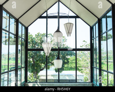 White hanging modern lamps decorated under the high roof in glass house cafe on green tree an sky background. Stock Photo