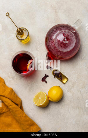 Transparency glass of hibiscus tea (karkade, Agua de flor de Jamaica) with lemon, glass teapot, honey with honey stick and orange napkin at white back Stock Photo